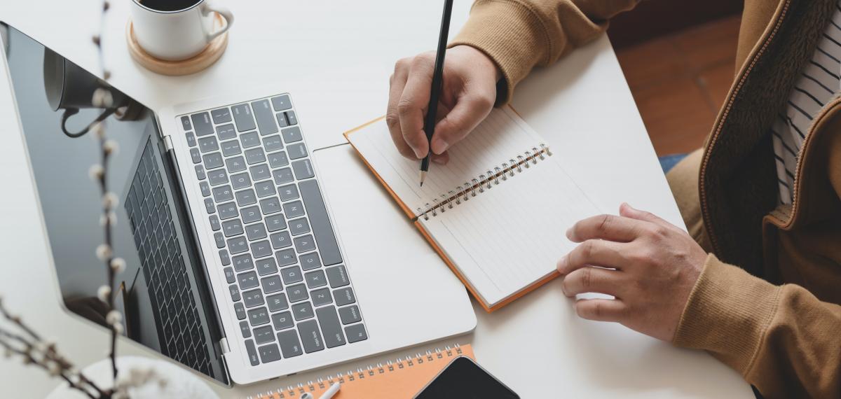 person-writing-on-notebook-next-to-laptop