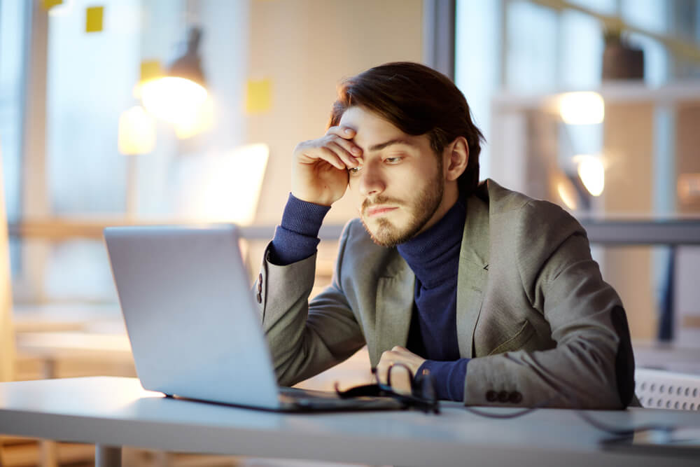 Tired Financial Manager Sitting at Desk and Finishing Annual Accounts With Help of Laptop, Interior of Dim Open Plan Office on Background