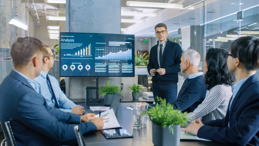 Young Stock Trader Shows to the Executive Managers Cryptocurrency and Trade Market Correlation Pointing at the Wall TV.